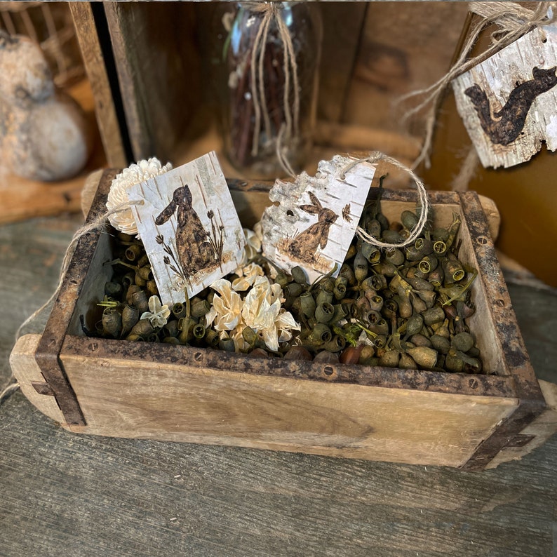 This shows a wooden bowl filler display. It has my handmade birch bark tags nestled in a wooden box of dried green botanicals. A decorative bundle of books has a fox tag tied to it.