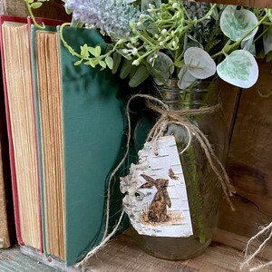 This image shows a mason jar flower arrangement with a birch bark accent tag tied around the rim. The tag has a bunny and butterfly wood-burned onto it.