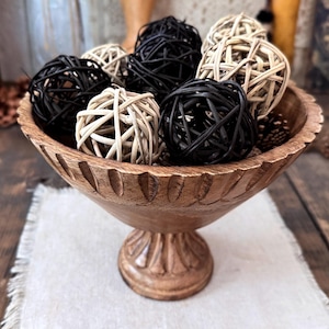 May include: A wooden pedestal bowl filled with decorative woven spheres in black and natural tones. The bowl has carved details and sits on a white cloth, creating a rustic, natural aesthetic. Pinecones are also visible.