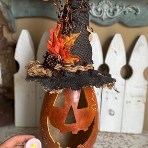 A Large handmade pumpkin witch used for fall decorating. Made from a real dried gourd. The pumpkin, named Hocus Pocus, has a tall burlap crinkled witch hat with a fall leaf arrangement. The pumpkin includes a battery-operated candle.