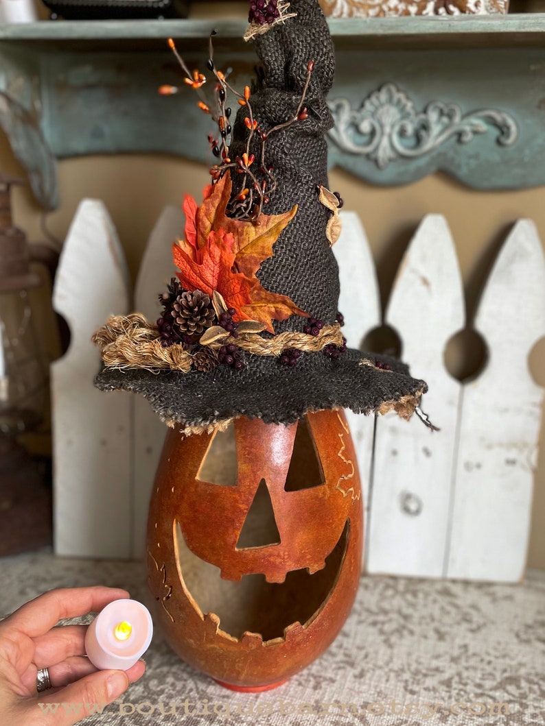 A Large handmade pumpkin witch used for fall decorating. Made from a real dried gourd. The pumpkin, named Hocus Pocus, has a tall burlap crinkled witch hat with a fall leaf arrangement. The pumpkin includes a battery-operated candle.