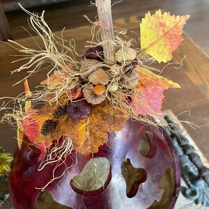This image shows a close-up of the Halloween gourd top. It has an arrangement of fall leaves, pinecones, and dried berries on top. The pumpkin gourd is sitting on a dinning room table.