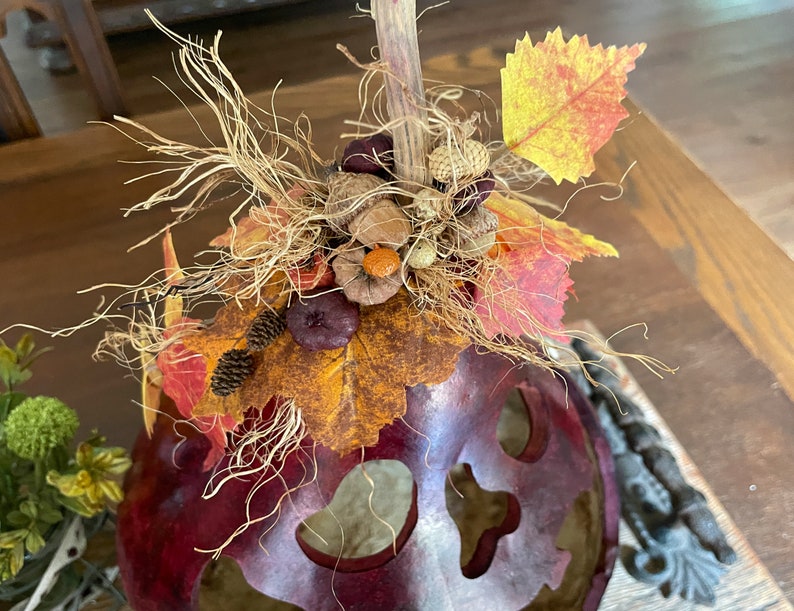 This image shows a close-up of the Halloween gourd top. It has an arrangement of fall leaves, pinecones, and dried berries on top. The pumpkin gourd is sitting on a dinning room table.