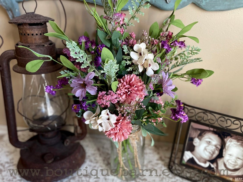 This image shows an artificial bouquet of flowers in a mason jar vase. It features purple daisies, pink mums, and white hydrangeas.