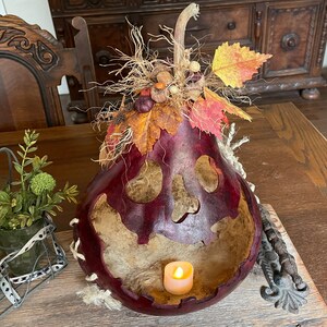 Pu&ograve; includere: Una zucca svuotata di colore rosso scuro che funge da lanterna, con un volto a teschio intagliato e un lumino acceso all'interno. La parte superiore &egrave; decorata con rafia secca, piccole ghiande, pigne e foglie autunnali artificiali.