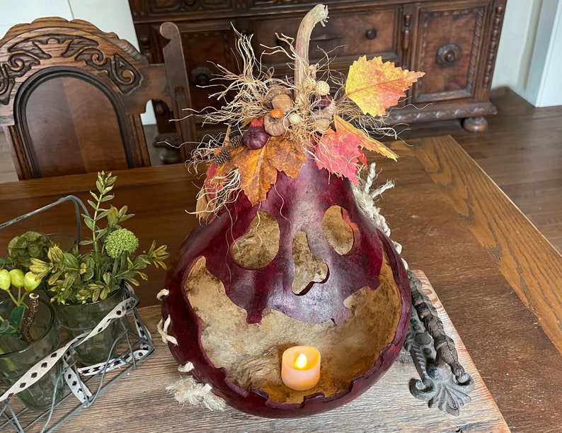 Pu&ograve; includere: Una zucca svuotata di colore rosso scuro che funge da lanterna, con un volto a teschio intagliato e un lumino acceso all'interno. La parte superiore &egrave; decorata con rafia secca, piccole ghiande, pigne e foglie autunnali artificiali.