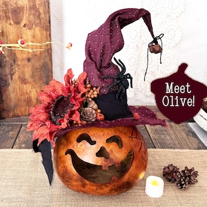 Può includere: Una zucca intagliata a forma di lanterna con un volto sorridente, che indossa un alto cappello da strega in tessuto viola scuro decorato con un grande fiore arancione, un ragno nero e piccole pigne. Il testo "Meet Olive!" è stampato su un cartello.