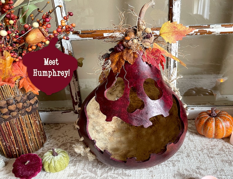 This image shows a large mouth pumpkin gourd decoration. It is a real dried gourd with a carved face. The face has a ghostly expression. The gourd is stained berry color. It has dried botanical berries and fall leaves arranged on the top.