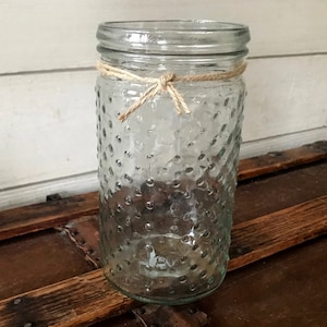 Op de afbeelding: Een glazen pot met een transparante, getextureerde oppervlakte en een bruine touw strik rond de bovenkant. De pot staat op een houten oppervlak.
