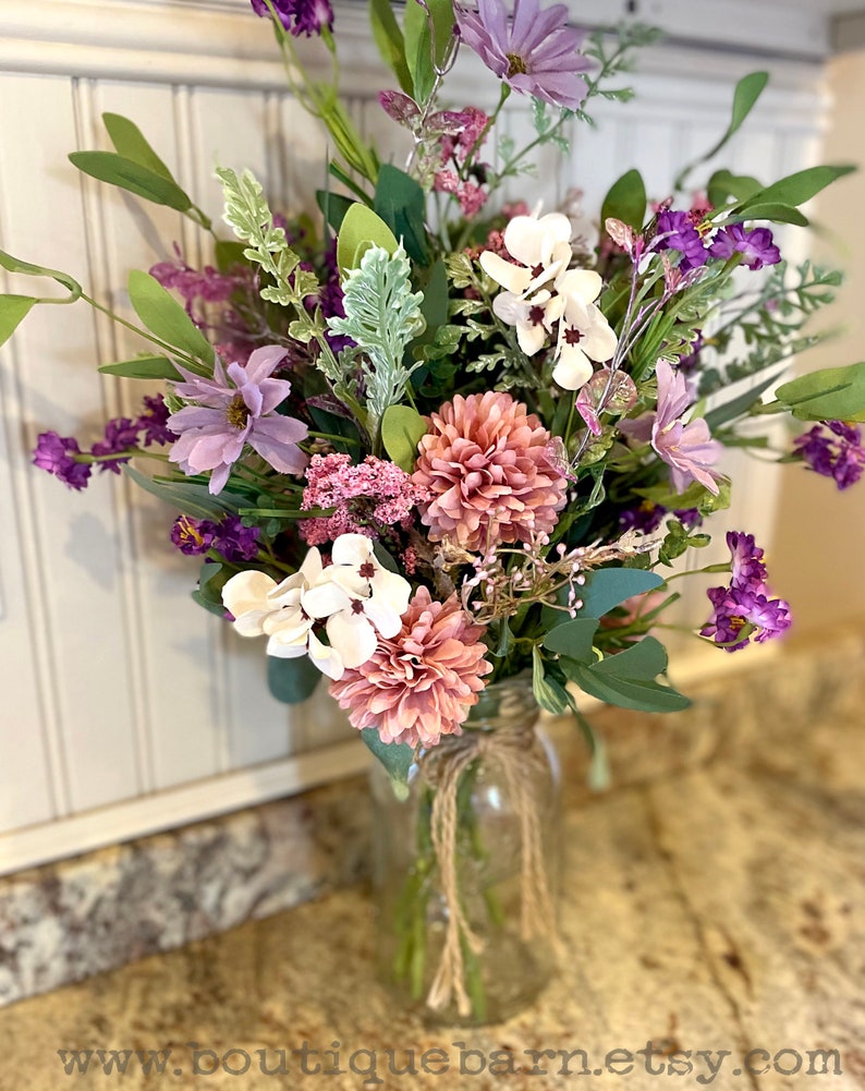 This image shows an artificial bouquet of flowers in a mason jar vase. It features purple daisies, pink mums, and white hydrangeas.