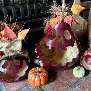 This shows three pumpkins sitting on a fireplace hearth. The pumpkins are made from real gourds. They are stained berry color. Each pumpkin has a ghostly expression carved for a face. The pumpkins are decorated with fall leaves and berries.