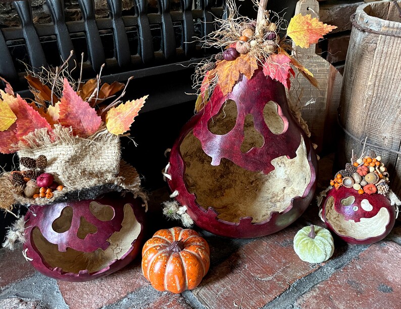 This shows three pumpkins sitting on a fireplace hearth. The pumpkins are made from real gourds. They are stained berry color. Each pumpkin has a ghostly expression carved for a face. The pumpkins are decorated with fall leaves and berries.