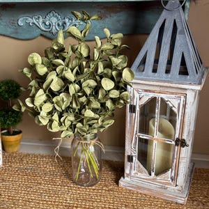 This shows a rustic entryway table display featuring a bouquet of green eucalyptus. The fake sprays are realistic looking arranged in a clear glass jar used as a vase. A decorative lantern with a candle sits next to the arrangment.