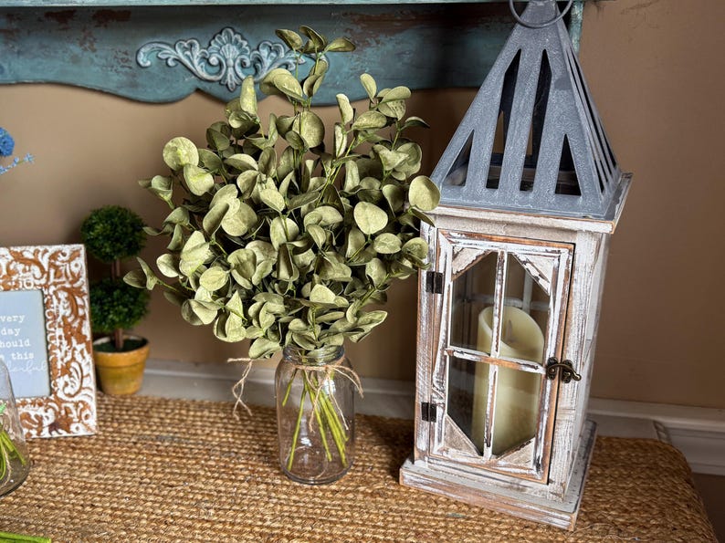 This shows a rustic entryway table display featuring a bouquet of green eucalyptus. The fake sprays are realistic looking arranged in a clear glass jar used as a vase. A decorative lantern with a candle sits next to the arrangment.