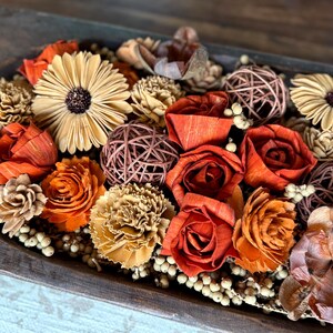 This shows one end of a long wood dough bowl with and arrangement of wood flowers and natural dried botanical berries. The flowers include roses, carnations, peonies and Gerba daisies. The display sits on a coffee table.