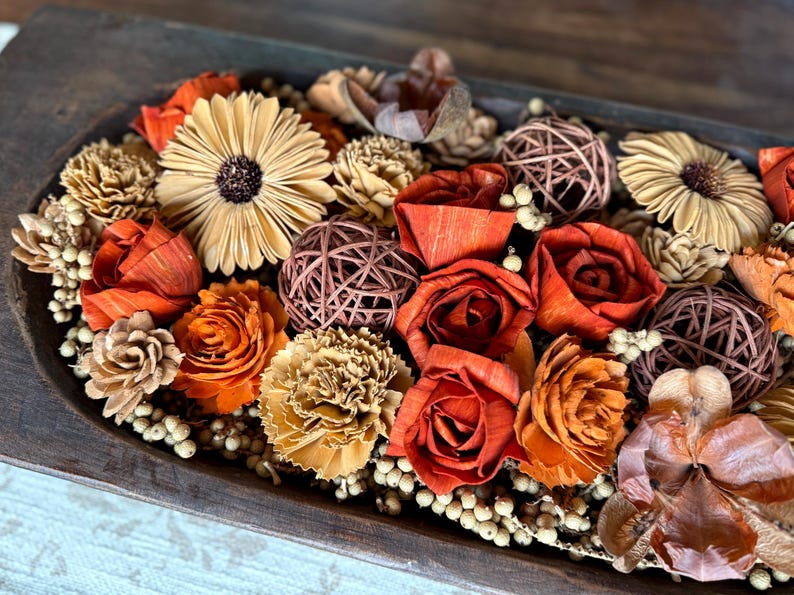 This shows one end of a long wood dough bowl with and arrangement of wood flowers and natural dried botanical berries. The flowers include roses, carnations, peonies and Gerba daisies. The display sits on a coffee table.