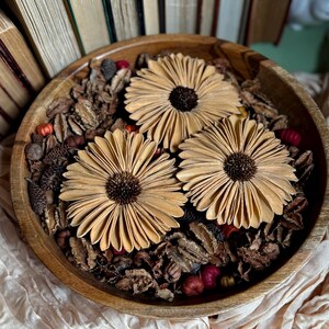 This shows three Gerba Daisy wood flowers arranged in a wooden bowl, sitting on an accent table. Several vintage books are standing in the background.