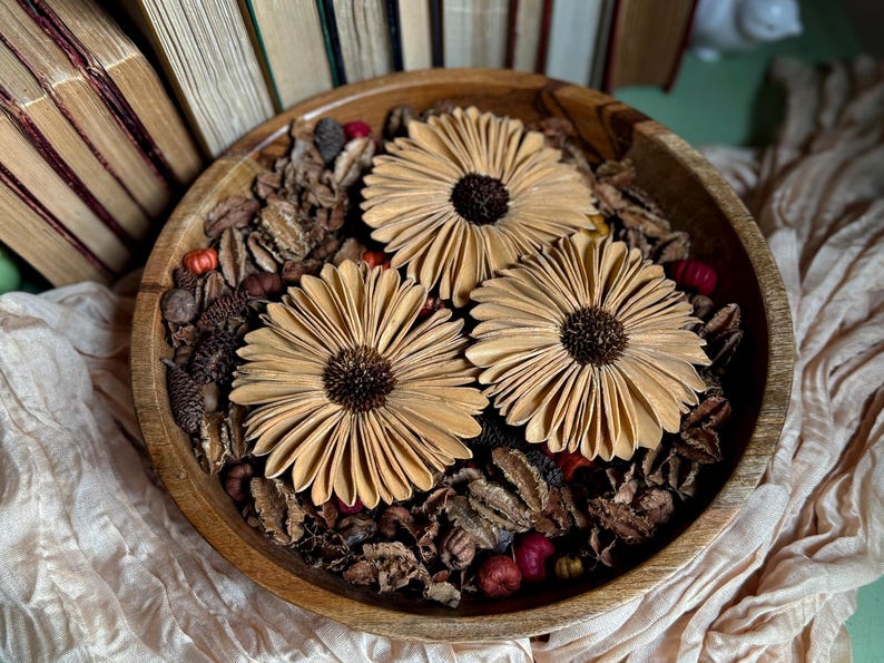 This shows three Gerba Daisy wood flowers arranged in a wooden bowl, sitting on an accent table. Several vintage books are standing in the background.