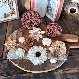 Puede incluir: Una tabla de servir de madera rústica exhibe una variedad de artículos decorativos. Estos incluyen esferas tejidas, flores de madera y adornos en forma de piña. La disposición se establece sobre un fondo de madera envejecida y otros elementos decorativos.