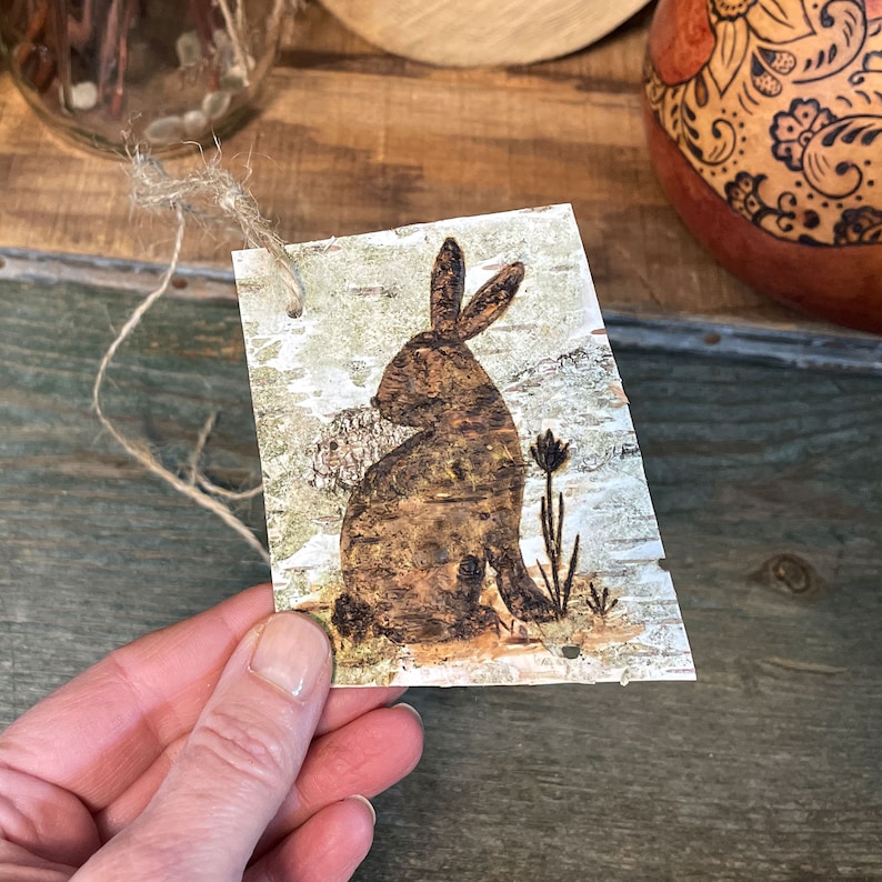 This shows me holding a birch bark tag. I hand-drew the rabbit artwork with pyrography tools. The bark tag has a hole punched in the corner with a piece of twine tied to it.