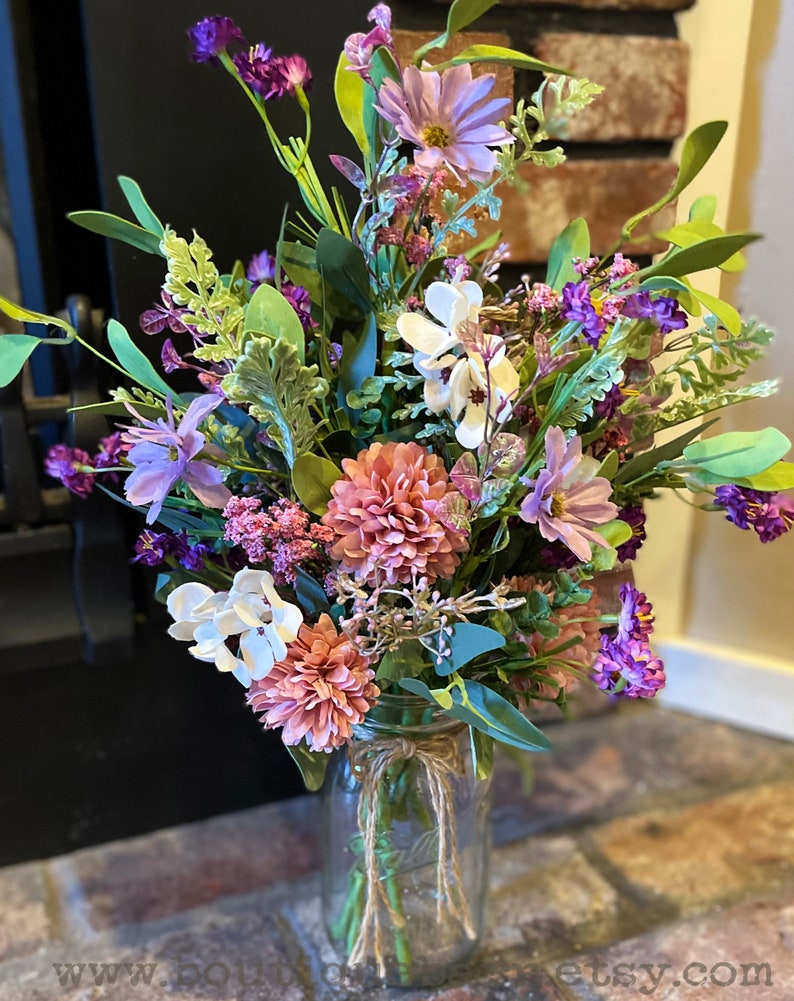 This image shows an artificial bouquet of flowers in a mason jar vase. It features purple daisies, pink mums, and white hydrangeas.