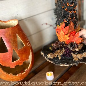 A Large handmade pumpkin witch used for fall decorating. Made from a real dried gourd. The pumpkin, named Hocus Pocus, has a tall burlap crinkled witch hat with a fall leaf arrangement. The pumpkin includes a battery-operated candle.