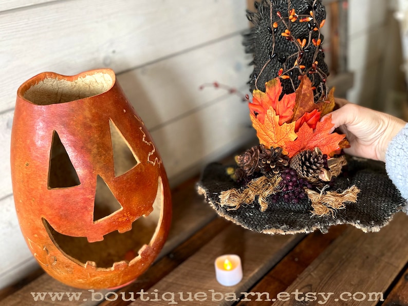 A Large handmade pumpkin witch used for fall decorating. Made from a real dried gourd. The pumpkin, named Hocus Pocus, has a tall burlap crinkled witch hat with a fall leaf arrangement. The pumpkin includes a battery-operated candle.