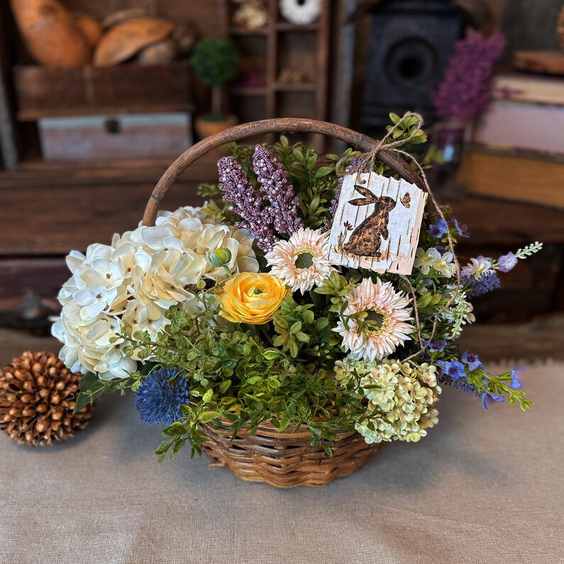 This shows a wicker basket full of faux flowers in white, green, and purple. My handmade birch bark tag with a bunny rabbit drawing is tied to the top of the basket