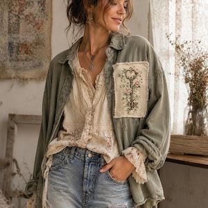Pode incluir: Uma camisa de botões verde-oliva com um emblema retangular no peito apresentando uma chave vintage e bordados florais. A camisa é usada sobre uma blusa creme com detalhes em renda e calças jeans azul-claro desgastadas com bordas desfiadas.