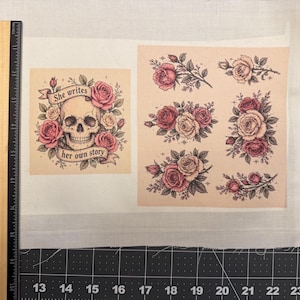 May include: A fabric sheet with printed designs. One design features a skull surrounded by pink roses, with the text "She writes her own story." The other designs are clusters of pink roses. A ruler is on the left side, with measurements in inches.