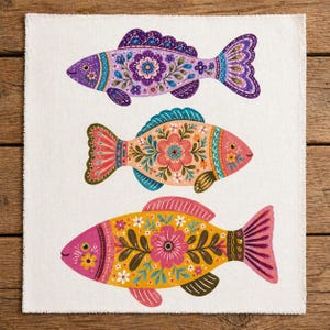 May include: A white rectangular decorative cloth featuring three colorful fish illustrations. Each fish is adorned with floral patterns in shades of purple, pink, orange, yellow, and teal. The fish have detailed fins and scales.
