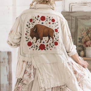 Könnte beinhalten: Eine beige langärmelige Jacke mit einem großen rechteckigen Aufnäher auf der Rückseite. Der Aufnäher zeigt einen braunen Bison in einem kreisförmigen Kranz aus roten Rosen und kleinen blauen Sternen. Die Jacke hat eine strukturierte Optik.