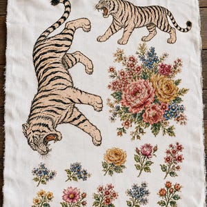 May include: A rectangular piece of off-white fabric with frayed edges featuring a large tiger in a downward pouncing pose, a smaller tiger walking, and a central bouquet of pink, yellow, and blue flowers, with smaller floral sprigs below.