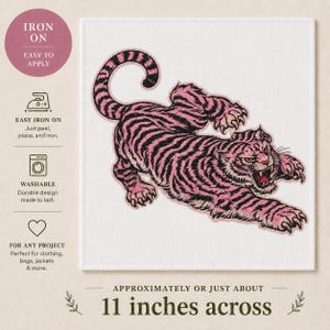 May include: An iron-on patch with a pink and black tiger design. The tiger is depicted in a dynamic pose, with its mouth open and claws extended. The patch is approximately 28 cm across.