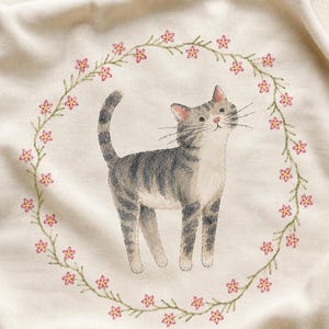 May include: A cream-colored fabric featuring a central illustration of a grey tabby cat with dark stripes, standing and looking upward. The cat is encircled by a delicate, hand-drawn wreath of thin green vines and small, five-petaled pink flowers.