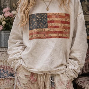 以下が含まれることがあります： 前面に色あせたアメリカ国旗のデザインが施された、明るいグレーのスウェットシャツ。星の青い部分と赤と白のストライプが特徴で、ヴィンテージ感があります。スウェットシャツは、花柄のパンツと組み合わせています。