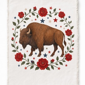 May include: A cream-colored woven throw blanket featuring a central illustration of a brown bison surrounded by a circular wreath of red roses with green leaves and scattered small red, white, and blue stars. The blanket has a fringed edge.