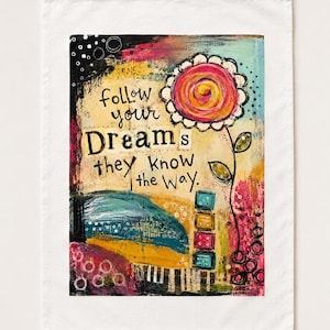 May include: A white rectangular tea towel featuring a colorful mixed-media illustration. The artwork includes a large stylized flower, stacked square blocks, and the text "follow your Dreams they know the way." The design uses black, pink, yellow, and blue.