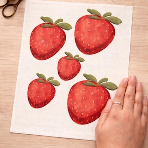 May include: A fabric with five watercolor-style strawberries. The strawberries are red with green leaves and small yellow seeds. A pair of scissors and a spool of green thread are in the upper left corner.