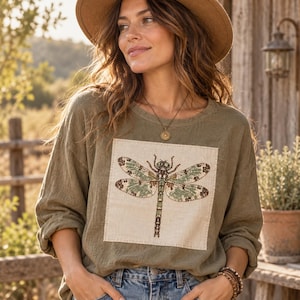 May include: An olive green long-sleeve shirt featuring a square cream-colored patch on the chest. The patch displays a detailed graphic illustration of a dragonfly with intricate wing patterns in shades of green, brown, and black.