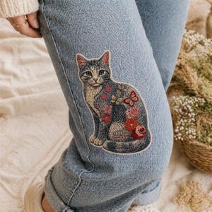 May include: Light blue denim jeans featuring an embroidered patch of a gray cat adorned with floral and butterfly designs in shades of red, pink, and blue. The patch is sewn onto the knee area of the jeans.