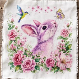 Peut inclure: Un panneau de tissu carré aux bords effilochés présentant une illustration imprimée d'un lapin violet entouré de roses roses et de feuilles vertes. Un colibri coloré vole à gauche et un papillon jaune et violet repose sur le nez du lapin.