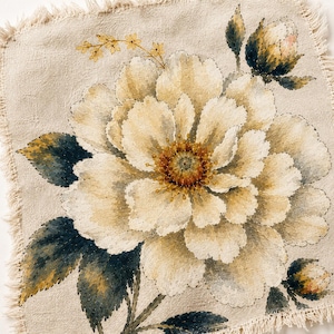 Puede incluir: Un textil cuadrado de lino beige con un diseño floral grande y detallado. La flor es principalmente de color crema con un centro amarillo y hojas de color verde oscuro. Los bordes del textil están deshilachados.
