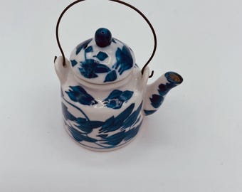 Puede incluir: Una tetera blanca pequeña con un diseño floral azul y un asa de alambre. La tetera tiene una tapa y un pico.