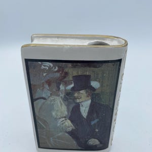 May include: White ceramic book-shaped decanter with a gold trim and a painting of a man and woman in a cafe scene.