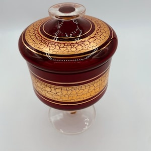 May include: A red glass jar with a gold rim and a gold crackle design. The jar has a clear glass lid and sits on a clear glass pedestal.