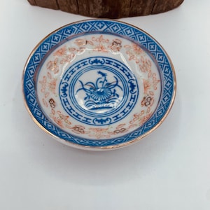May include: A small, white ceramic dish with a blue and gold rim. The dish features a floral design in the centre with a blue and white pattern around the edge.