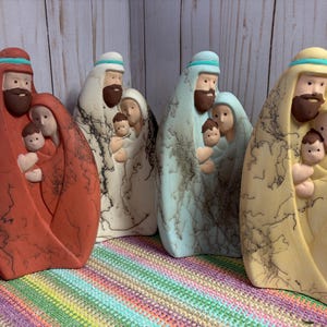 Puede incluir: Cuatro figuras de belén de cerámica en rojo, blanco, azul claro y amarillo. Cada escultura representa una familia de tres con una figura barbuda, una persona y un bebé, todas con un distintivo patrón de cocción negro estilo crin de caballo.