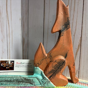 Puede incluir: Una escultura de cerámica color terracota de un coyote aullando, con marcas negras estilo crin de caballo. La base es de color verde azulado con un diseño de pluma negra. Una tarjeta de visita de Burnt Earth Ceramic Clay & Glass Art está insertada.