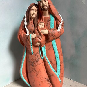 Puede incluir: Una figurita de cer&aacute;mica de la Sagrada Familia. Las figuras est&aacute;n pintadas en tonos marrones y rojos, con detalles turquesa. La madre sostiene un beb&eacute; en sus brazos. El padre est&aacute; de pie detr&aacute;s de ellos, con su brazo alrededor de la madre.